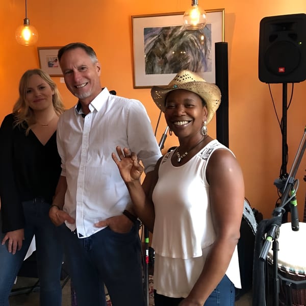 Three smiling people, two men and one woman, stand together in a restaurant with musical instruments.